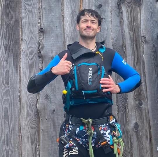 Person in a wetsuit giving thumbs up, standing against wooden wall.