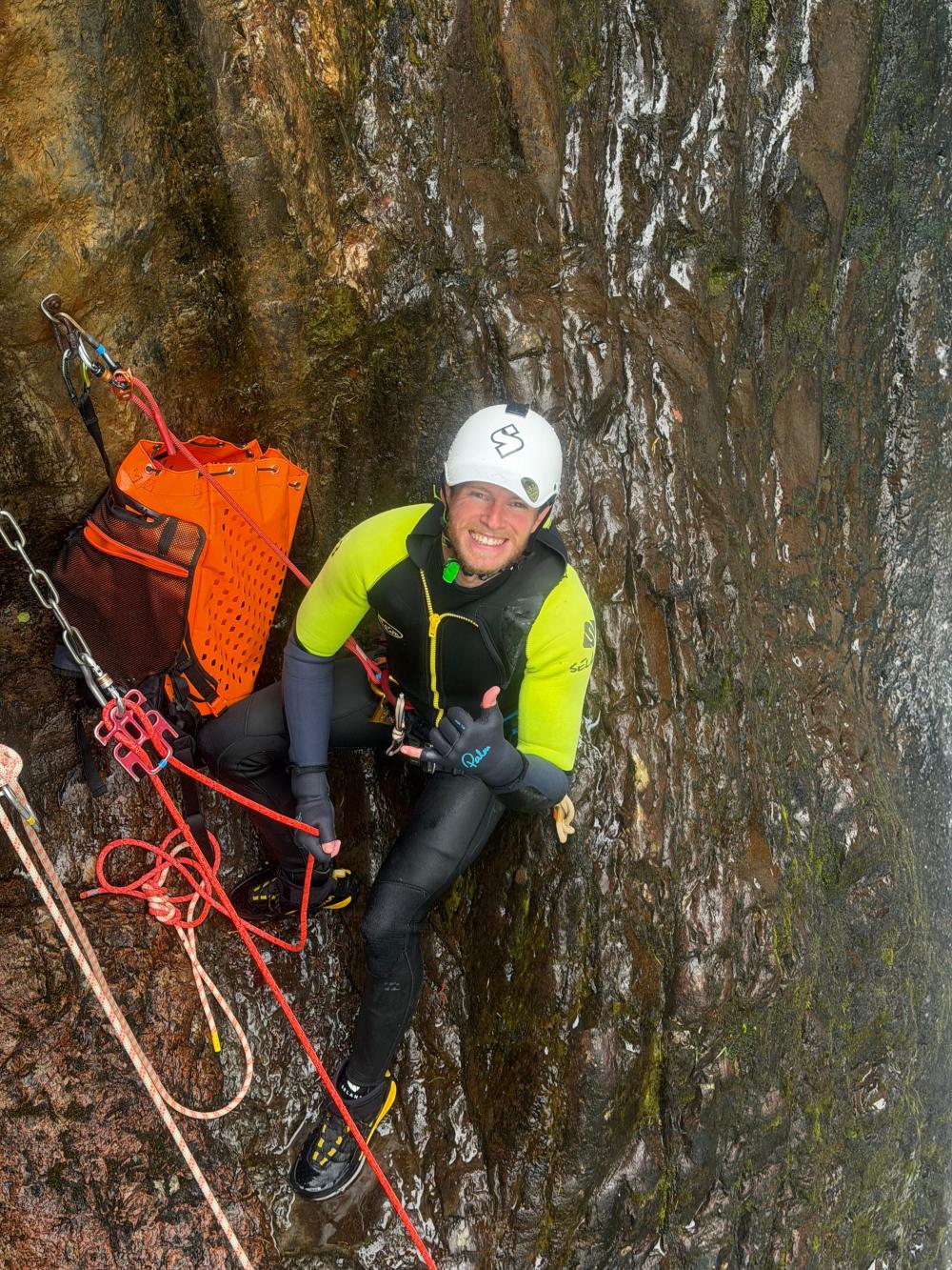 Rock climber with helmet gives thumb up while climbing a wet, steep wall with ropes and gear.