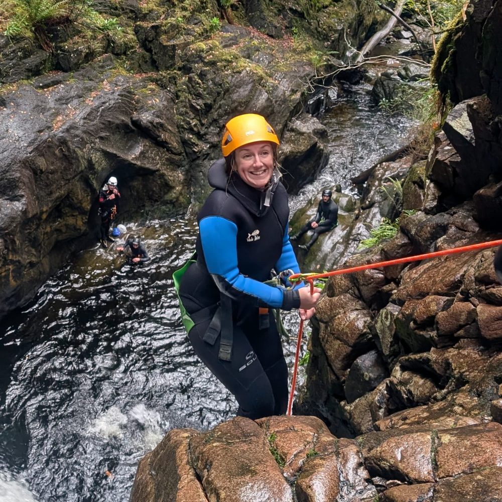Person in helmet abseiling down a rocky gorge near a waterfall.