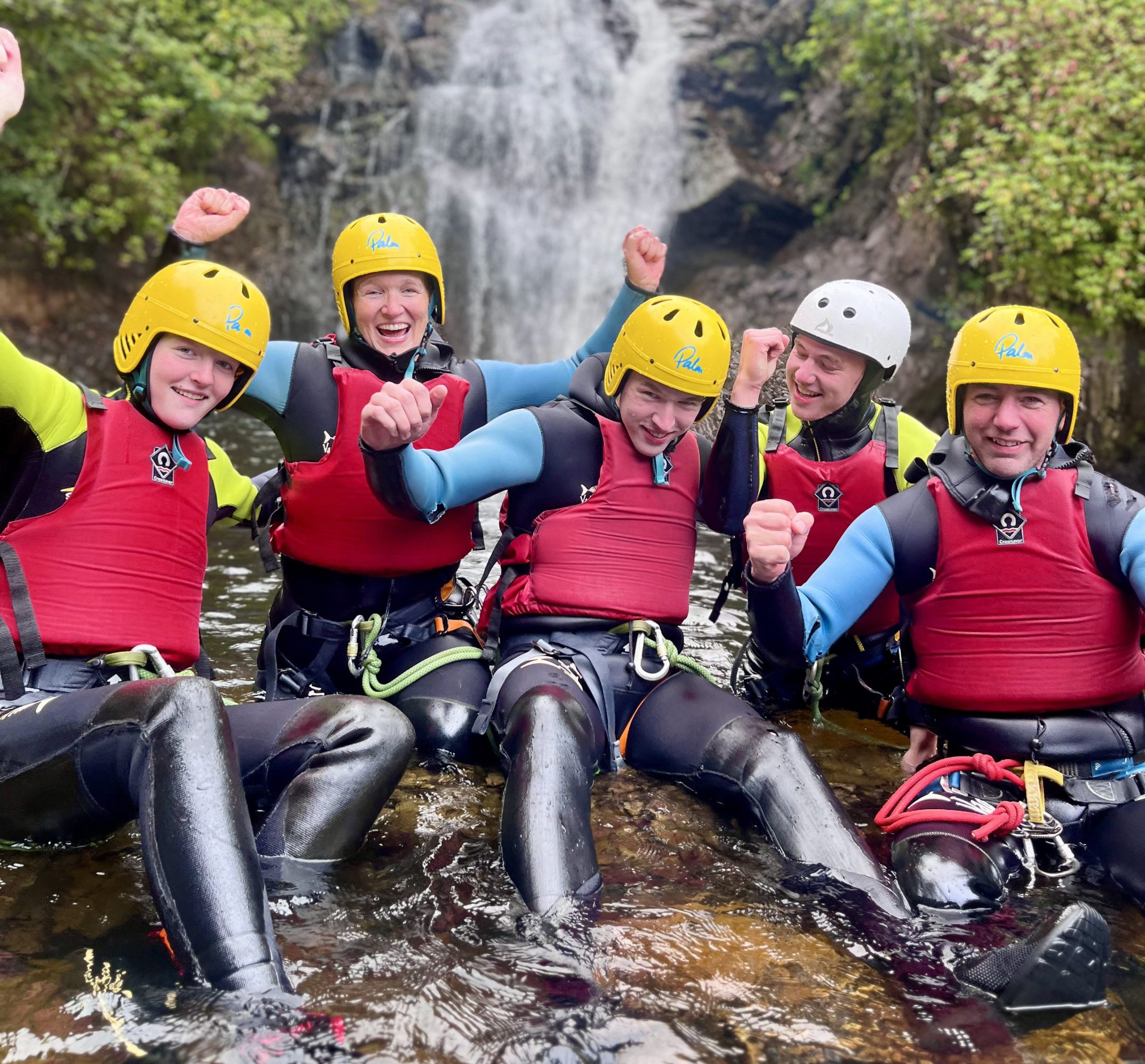 Group of people in wetsuits and helmets sitting by a waterfall.