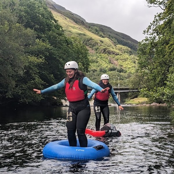 Two people in helmets standing on inflatable tubes in a river with greenery and hills around.